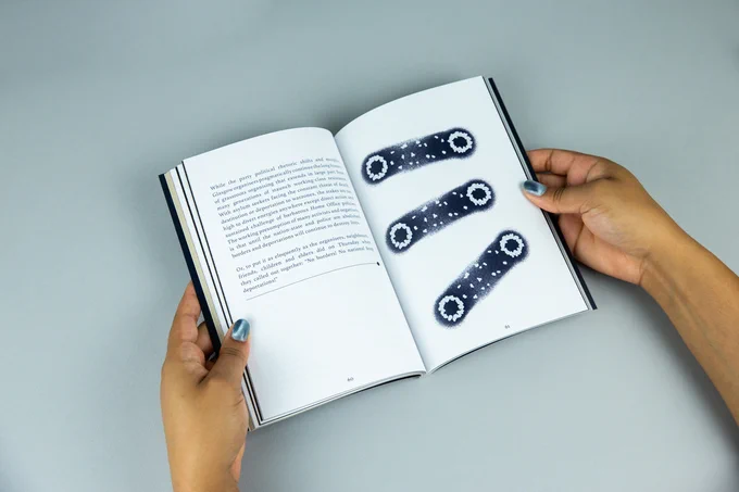 A small book being held open by a pair of hands with shimmering silver nail polish. The left has a column of text, some slogans jump out such as ​‘No borders! No nations! Stop deportations!’. The right page has an abstract drawing in white and deep navy, there seems to be six quivering white circles joined together in pairs by streaks of navy, the navy line is speckled with white dots like stars.