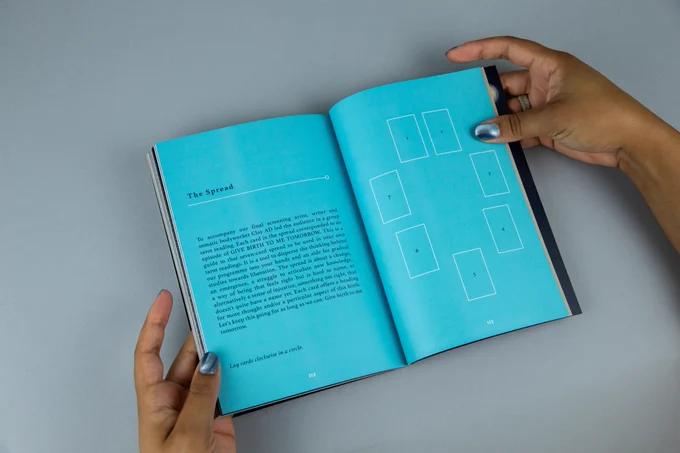 A pair of hands holds open an A5 size book. The interior is a bright cyan with white text. The hands holding the book have shimmering silver nail polish and wear a ring. The background is a light grey.