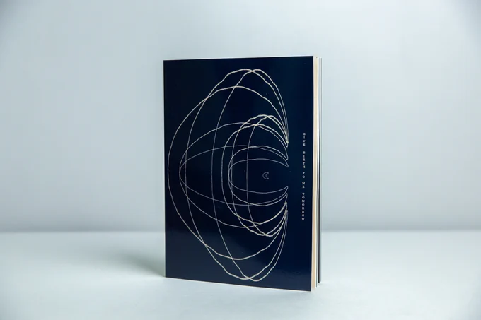 A small dark blue book stands upright on a white table against a white wall. A pale peach line illustration on the cover makes varying concentric circles around a small moon, and the words ​‘GIVE BIRTH TO ME TOMORROW’ are shown vertically on the right hand side.