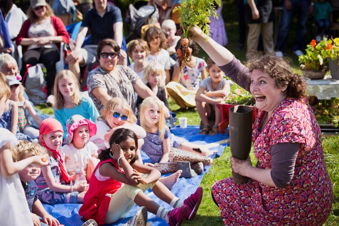 A lively outdoor scene with a woman in a floral dress enthusiastically holding up a bunch of freshly harvested carrots to a group of seated children. The children, along with some adults in the background, are sitting on blue tarps, watching with curiosity and excitement. The atmosphere is cheerful, with the event taking place in a natural, green environment that highlights a hands-on approach to learning about gardening and sustainability.