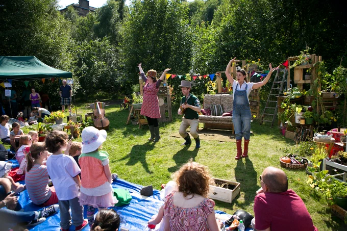 A vibrant outdoor scene featuring a large group of people, including children and adults, gathered on a grassy area in front of a green tent. The audience is seated on blue tarps, attentively watching two performers, a man and a woman, who are animatedly interacting with the crowd. The setting includes various plants, small gardening tools, and a rustic, eco-friendly atmosphere, reflecting a community event centred around sustainable practices.