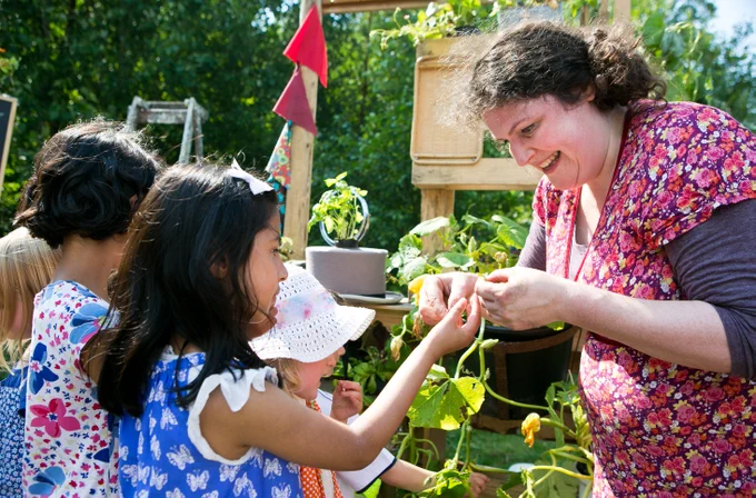 A close-up of a woman wearing a floral dress, joyfully engaging with a group of young children in a garden setting. The woman is handing a plant leaf to one of the children, who are eagerly watching and participating. The backdrop includes lush green plants and garden elements, emphasizing the educational and interactive nature of the event, which focuses on gardening and environmental awareness.