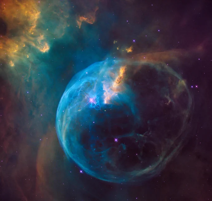 The Bubble Nebula. A glowing, blue, and roughly spherical structure surrounds pink stars in a hazy cosmic vista full of green, yellow, and gold dust and gas.