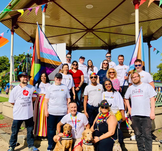 A large group of colleagues (and two dogs) posing for a picture at the bandstand in Leamington Spa, at Warwickshire Pride 2024.  