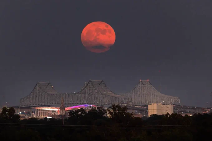 A "super blue moon," which appears in this photo as a hazy shade of red, rises above the Mississippi River in New Orleans on Aug. 30, 2023. A three-pointed bridge is visible below. Image credit: NASA/Michael Democker