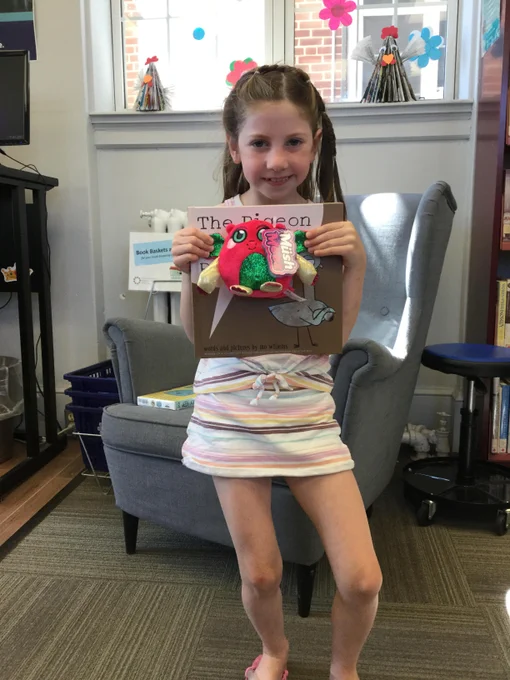 A young girl with long brown hair in braids, smiling, wearing a white summer striped dress, and holding up a Mo Willem Book, "This Pidgeon Needs a Bath!", and a cute stuffy animal. 