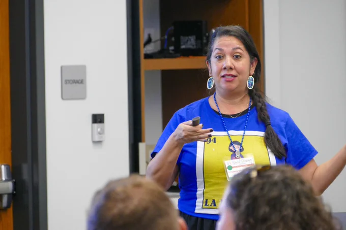 A person giving a presentation in a room wearing a blue shirt, standing in front of a technology shelf and gesturing to an audience.
