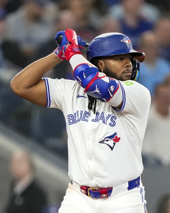 Vladimir Guerrero Jr. prepares to swing a bat. He is wearing a white Toronto Blue Jays jersey with blue lettering and blue Blue Jays batting helmet.