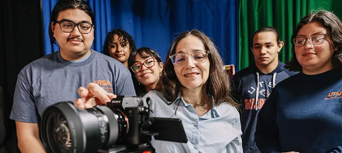 Group of five UTSA students and a professor engaging with a video camera in a studio setting.