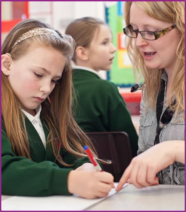 Photograph of young girl with a hearing aid in a school classroom writing on a notepad, with her teacher sitting next to her.