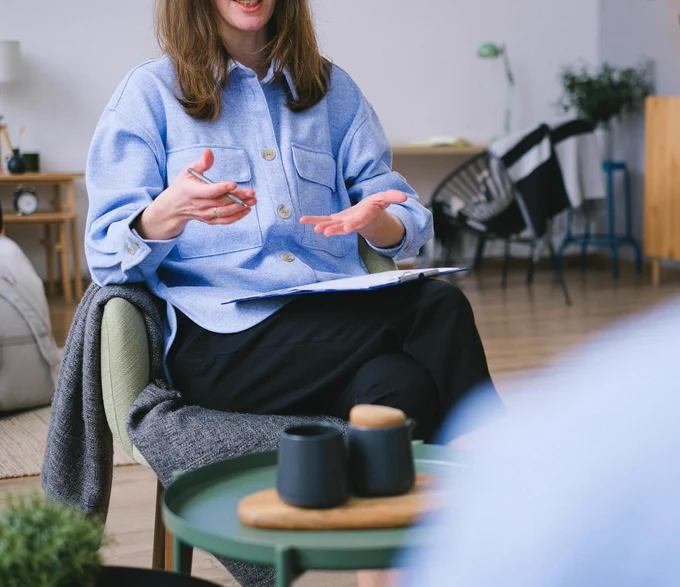 A therapist is sitting in a chair, smiling and gesturing to an off-camera client. 