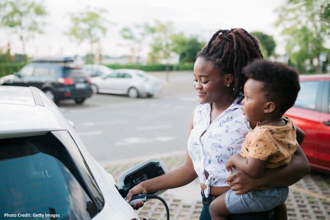 Adult charging an electric car while holding a toddler in a parking lot. Photo Credit: Getty Images.