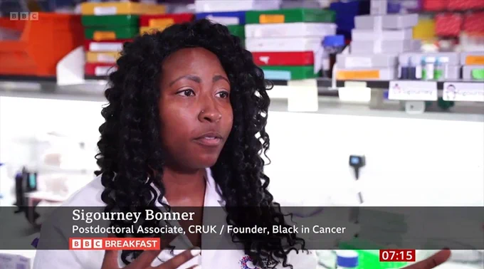 A screenshot of BBC Breakfast. Sigourney is in a lab wearing a lab coat. The on-screen banner reads: "Sigourney Bonner. Postdoctoral Associate, CRUK / Founder, Black in Cancer".