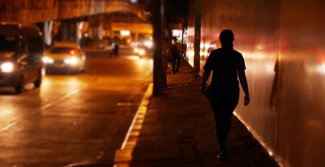 Donna cammina da sola su un marciapiede di notte mentre le macchine passano accanto a lei - App per la sicurezza