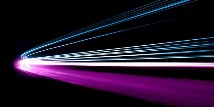 Night-time photo of a vehicle speeding around a bend with light trails captured above the road, creating a dynamic effect. The image is taken with a high shutter speed, emphasising the movement and energy of the scene.
