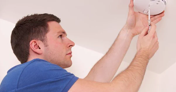 Image of man wearing a blue t shirt and he is fixing a smoke detector, holding a screwdriver next to the white detector on the white ceiling. 