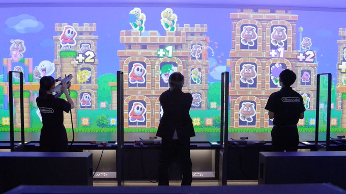 Screenshot from the Nintendo Museum Direct, with Shigeru Miyamoto and two museum employees playing the Zapper and Super Scope game. Cut-outs of Super Mario enemies are being hit with colourful splats.