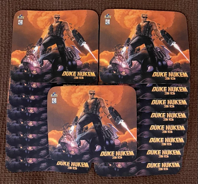 A collection of Duke Nukem 3D mouse pads featuring the iconic artwork of Duke standing triumphantly on top of an alien’s head with explosions in the background. The mouse pads are laid out neatly in a grid, showcasing the classic 3D Realms logo and the ‘Duke Nukem 3D’ title on each pad.