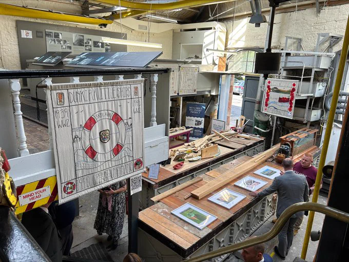 Image of workshop taken from a high vantage point. A banner is hanging with an image of a life ring on it. Two men are chatting in front of a bench which contains a row of four framed pictures (detail cannot be seen).