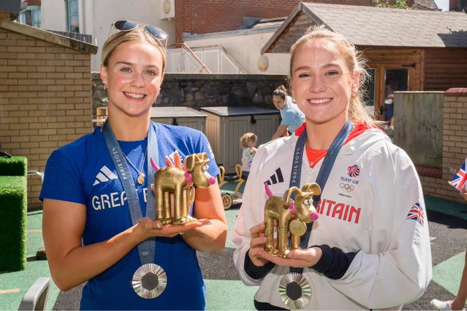 Kate and Izzy are standing in the playground. They are both wearing their Olympic silver medals around their necks and are holding up their Golden Gromit statues.