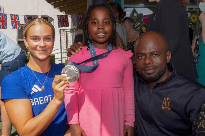 A young girl is wearing Kate's silver medal around her neck. Kate is standing next to her on her left and the girl's dad is on her right. 