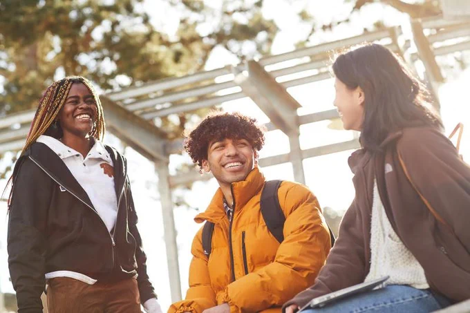 Three students smiling and engaging in a lively conversation on Malcolm X Plaza on a sunny day.