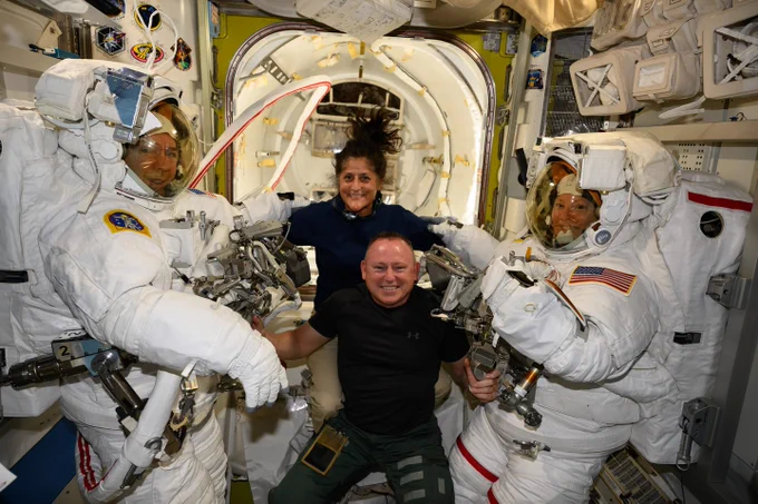 NASA astronauts Suni Williams and Butch Wilmore, wearing dark shirts and long pants, grin at the camera as they float inside the International Space Station. Two astronauts wearing full spacesuits, in preparation for a spacewalk, are on Butch and Suni's left and right. Credit: NASA