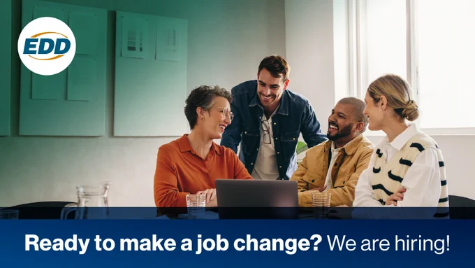 Four people gathered around a laptop in a brightly-lit office, smiling and engaged in a discussion, with a poster on the wall featuring the logo of the Employment Development Department (EDD) and text stating "Ready to make a job change? We are hiring!