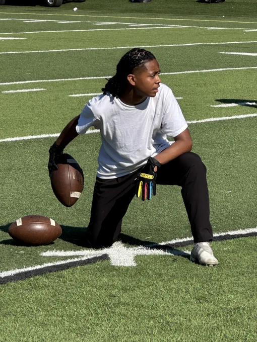 A Lincoln High School student-athlete is kneeling on the football field while holding a football; he is wearing a white t-shirt, black pants and a black glove with yellow, red and blue fingers