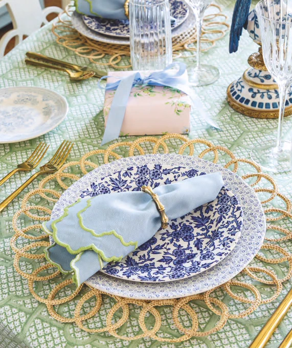 First: A place setting with a green lattice-print tablecloth, straw place mat, the Burgess & Leigh Felicity Blue dinner plate and Arden Blue salad plate, blue napkins embroidered with a green scalloped edge from Mrs. Alice tucked in a gold napkin ring from Blue Pheasant, gold flatware, Vietri glassware, and a petite gift wrapped in paper from Simply Jessica Marie. 

