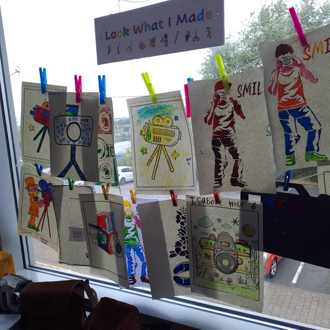A sign that reads, 'Look What I Made' hanging on a window with many sheets showing drawings and colourings in of cameras and people taking photos 