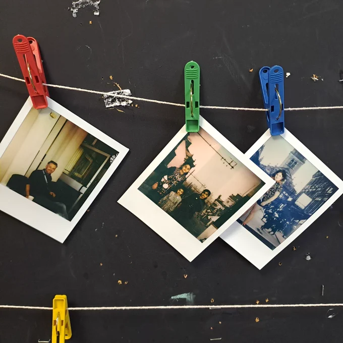 Three polaroids hanging from pegs on a blackboard showing people posing in front of a historic photo of Nelson