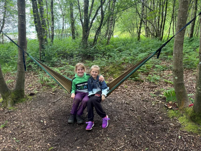 two girls sat in a hammock smiling with their arms round one anothers shoulders
