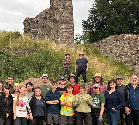 A group of smiling people stand at the bottom of a grassy bank looking at the camera.  At the top of the bank are the ruins of the castle. 
