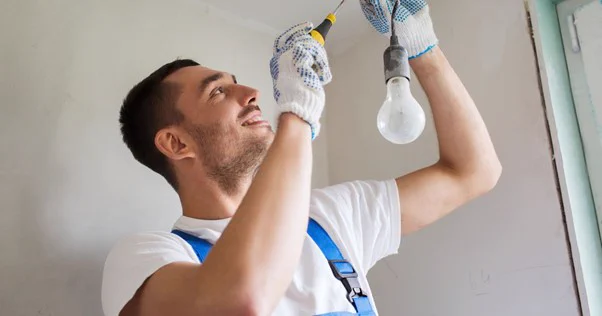 Image of man wearing workman dungarees and white t shirt. He is holding a screwdriver, wearing gloves, and holding a wire with a lightbulb attached to the bottom. He is smiling.