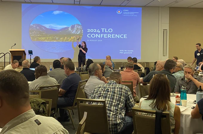 A speaker stands on stage with a slide displayed behind her with the words 2024 TLO Conference and more than 100 participants sitting in front of her listening.
