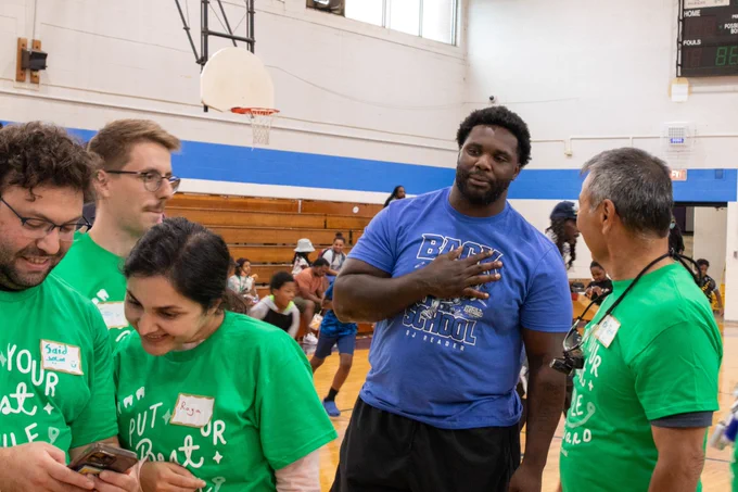 DJ Reader of the Detroit Lions stands in a blue shirt talking to TeamSmile volunteers wearing green shirts. 