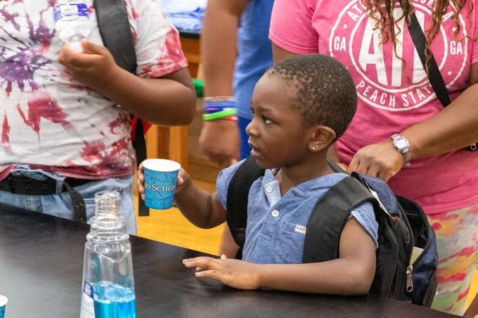 A young Black boy wearing a giant backpack holds a cup of mouthwash.