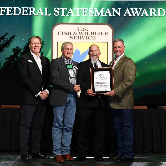 Four men in brow and black suits stand accepting an award, smiling at the camera. 