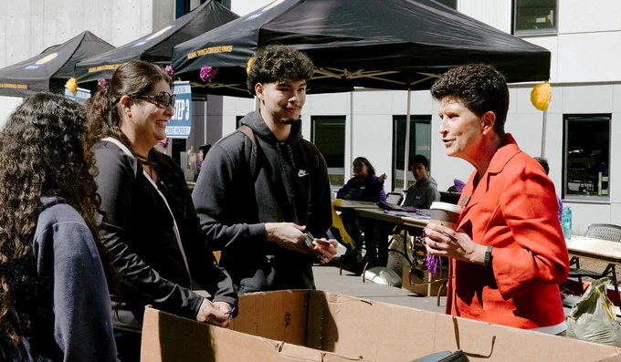 President Lynn Mahoney greets three family members outside of the West Grove Commons with others visible in the background on a sunny day. Mahoney is wearing a red blazer and holding a disposable coffee cup. The setting includes festive decorations and a busy, community environment.