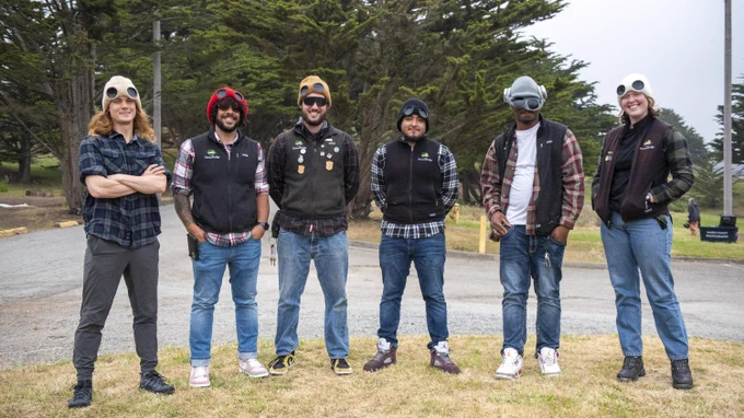 Six NatureBridge staff all wearing knit beanies and NatureBridge branded gear stand in a line outdoors and smile at the camera.