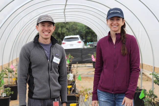 Two individuals with UC Cooperative Extension are standing inside a greenhouse, smiling at the camera, surrounded by potted plants.