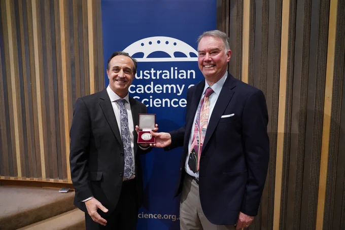 Two people stand smiling while holding a medal in a box. A banner of the Australian Academy of Science s behind them.