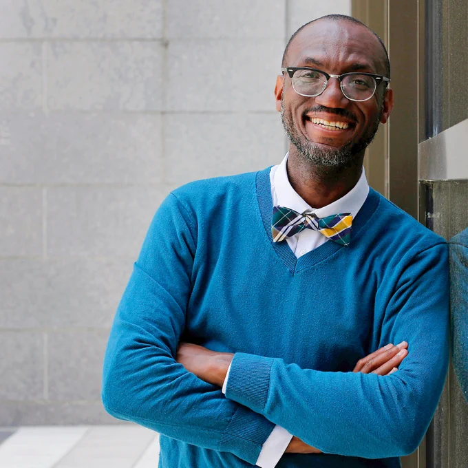 André Isaacs in a blue sweater and patterned bow tie leaning on a wall.