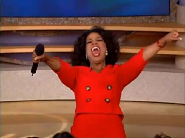 Oprah Winfrey raises her hands excitedly while wearing a red outfit.
