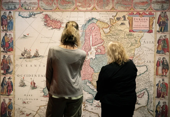 Two people stand in front of a wall-sized map of Europe from the Renaissance period. They are facing away from the camera, appearing to ponder the wall.
