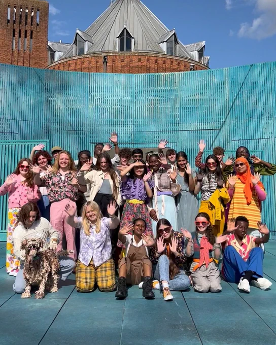 A group photo of the Next Generation Act company for The Two Gentlemen of Verona on the Garden Theatre stage. They are wearing colourful costumes and waving at the camera. The Swan Theatre is just visible behind the set against a bright blue sky.