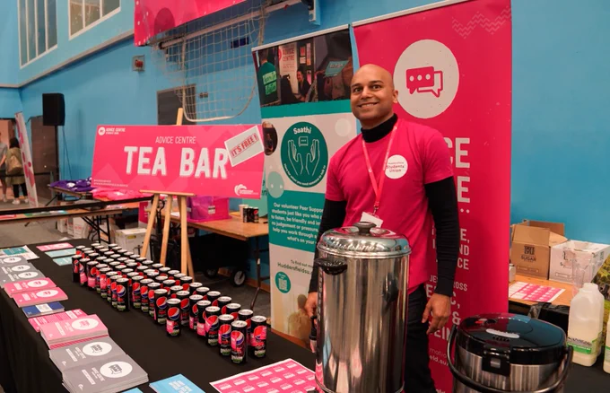 A person standing behind a "Tea Bar" at a health event booth, smiling at the camera with promotional materials and beverages on display.