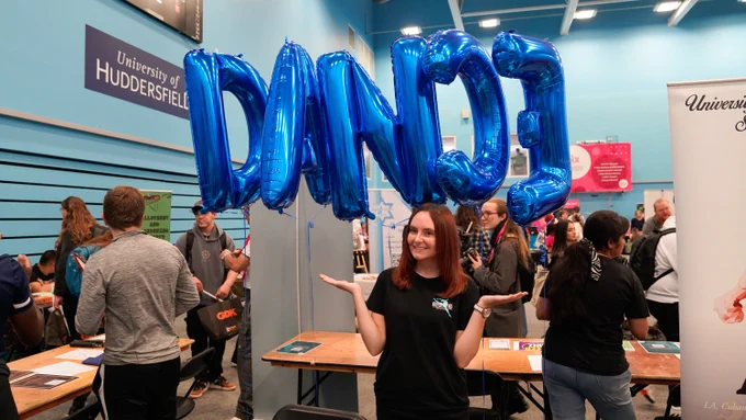 A person holding up blue letter balloons spelling "DANCE" at a University of Huddersfield event.