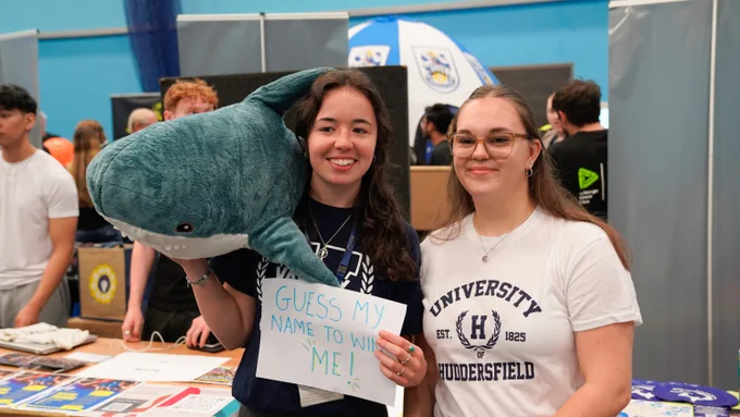 Two individuals at a promotional booth, one holding a sign that reads "Guess my name to win me!" alongside a large plush shark. Behind them are banners related to the University of Huddersfield.